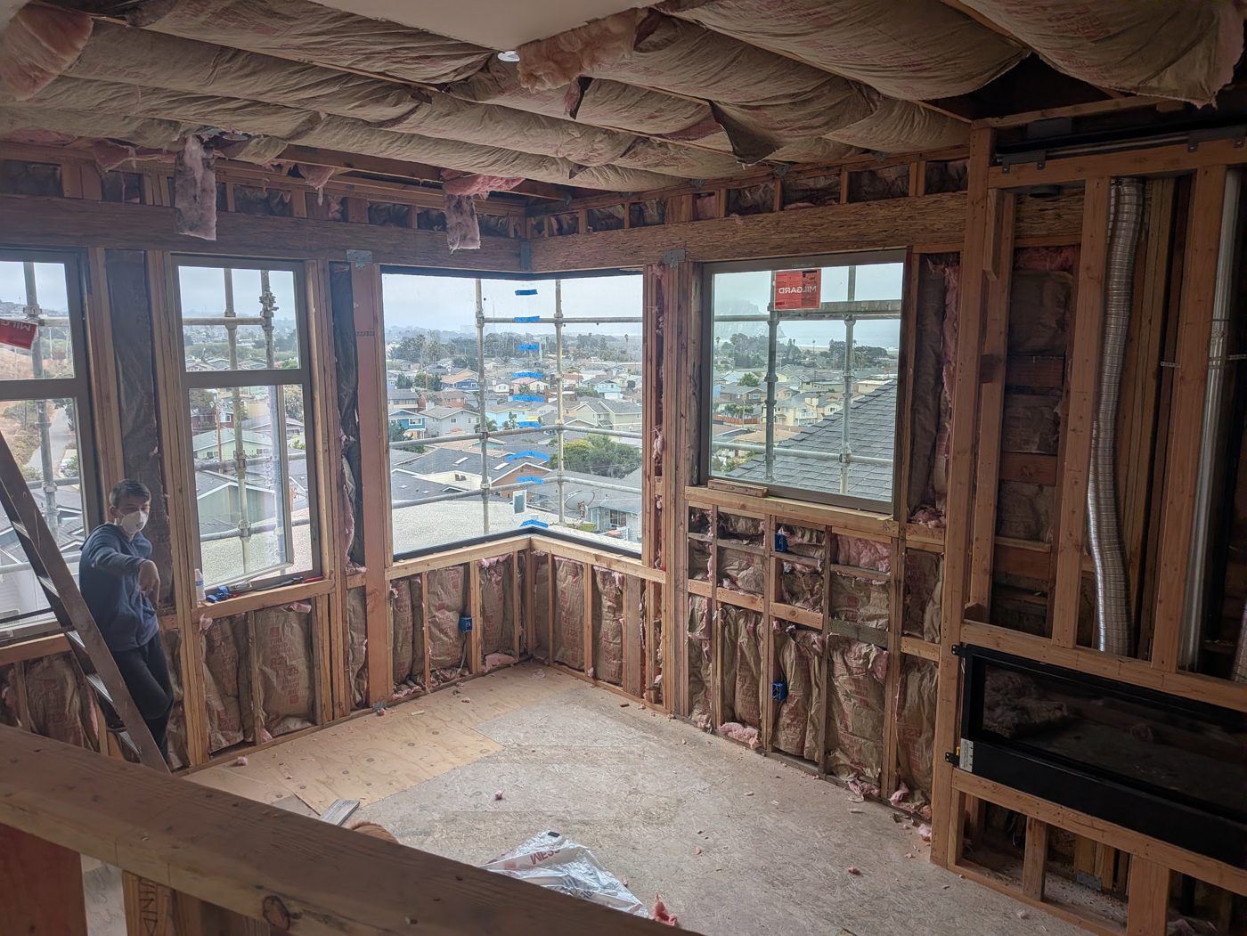 Wide view of the corner-window room with insulation batts in the walls and ceiling, a worker on the left