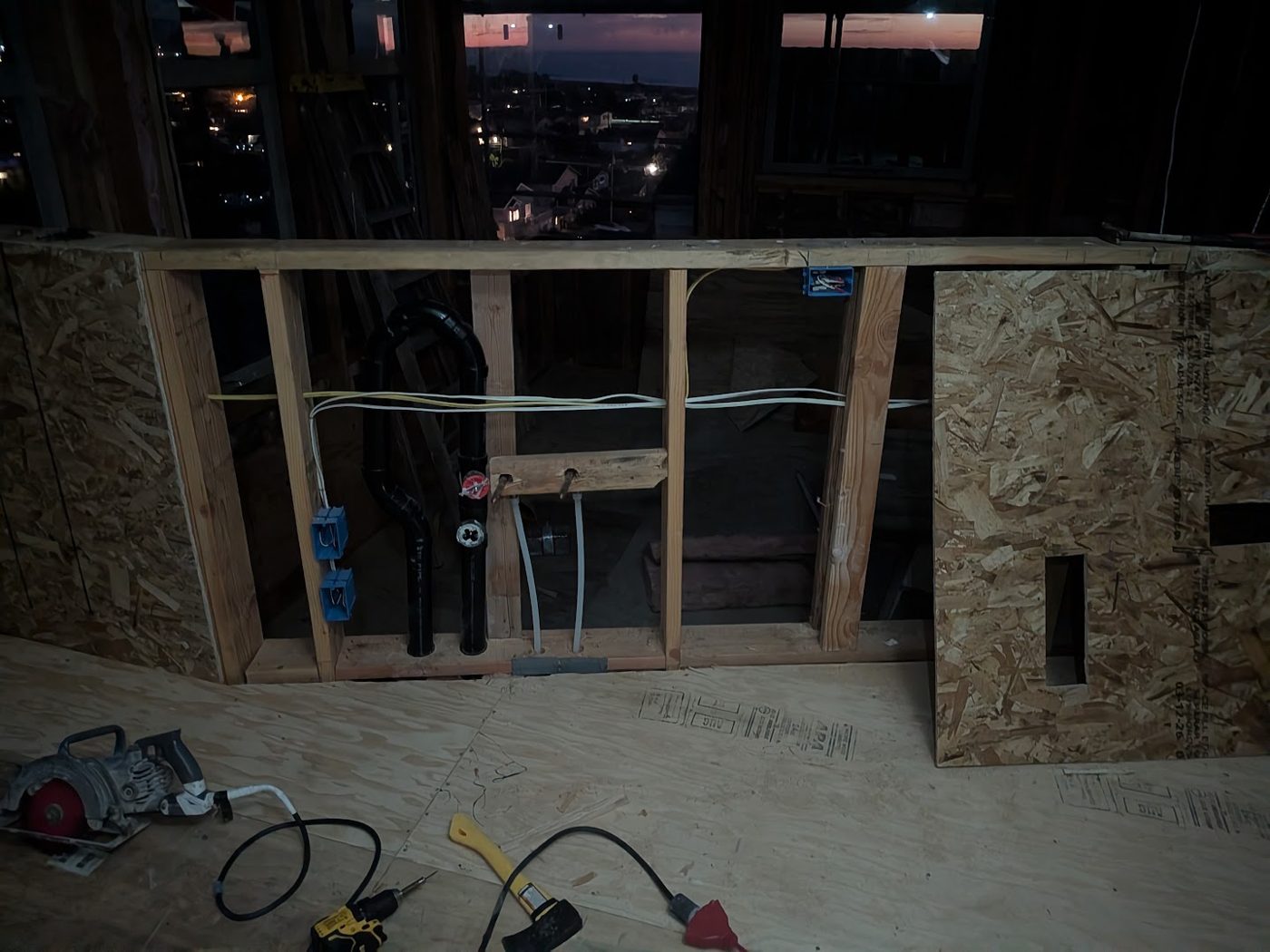 Open wall framing at dusk with drain line, water supply lines, wiring, and outlet boxes roughed in, Morro Bay lights through the windows
